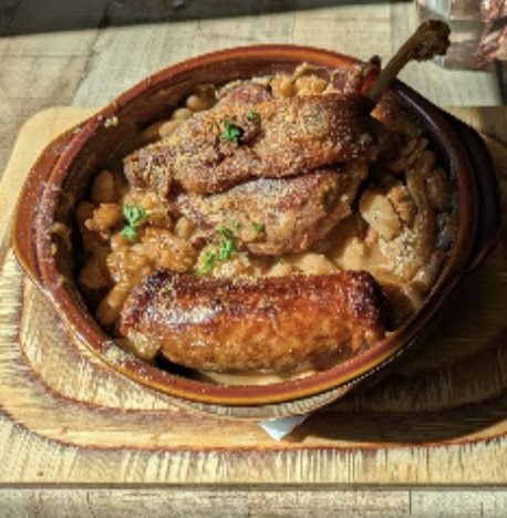 Confit et saucisse dorés dans une cassole en terre cuite, posée sur une planche de bois.