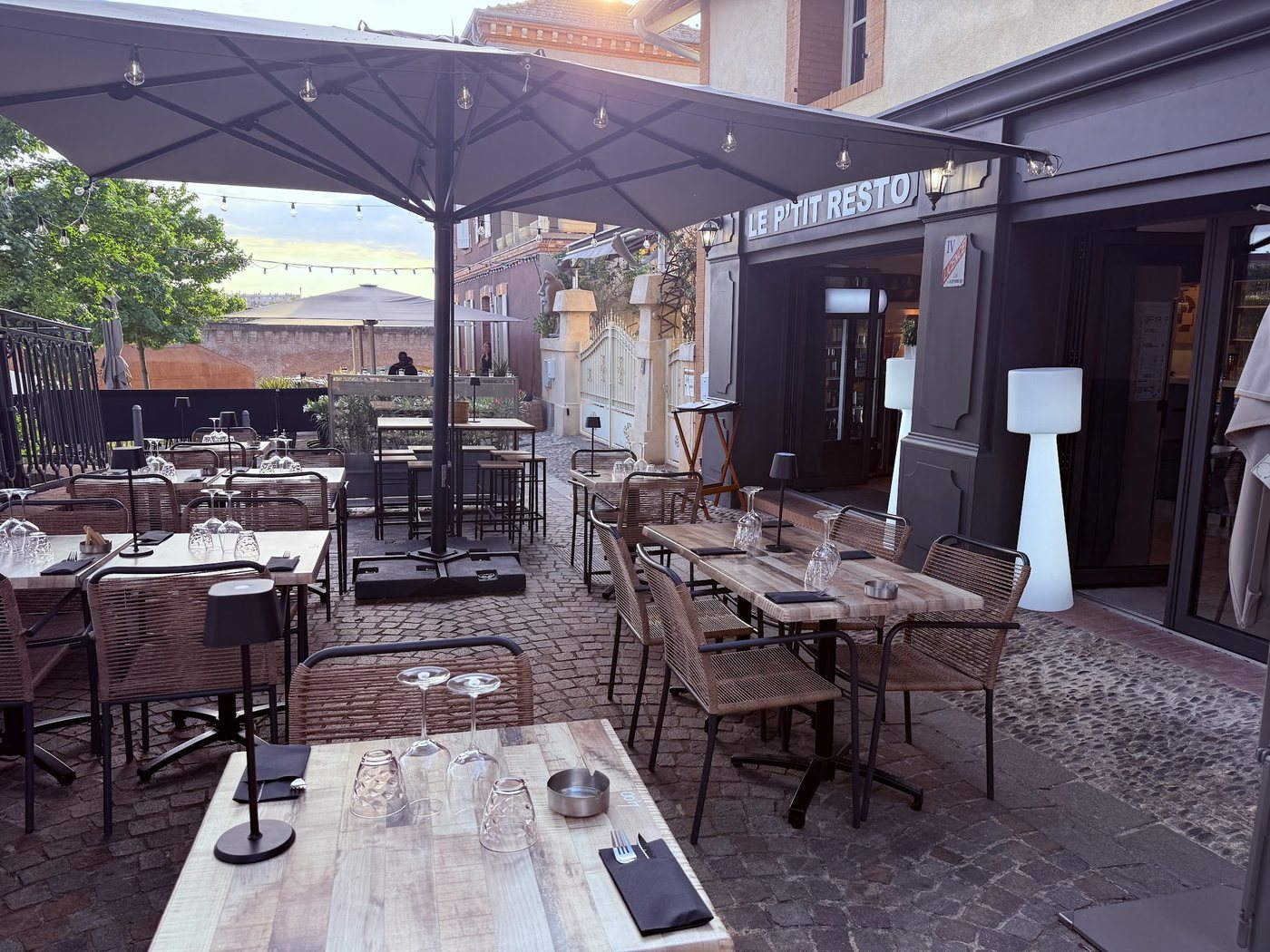 La terrasse du P’tit Resto sous un grand parasol gris, guirlande de lampes, tables dressées et chaises en rotin sur les pavés, devant l’enseigne.